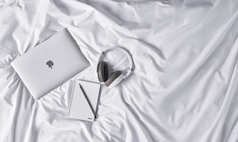 white headset on white bed sheets next to a mac laptop and a white notebook