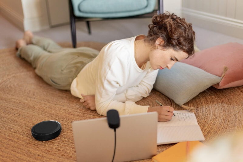 Woman working from home on laptop, writing in notebook with Jabra Speak2 75 on the floor